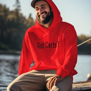May include: A man wearing a red hooded sweatshirt with the text "Got Certs?" in black lettering. He is sitting on a wooden dock with a body of water in the background.