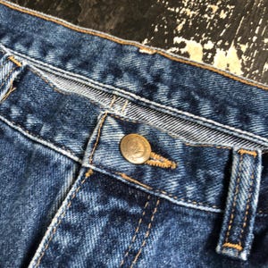 Op de afbeelding: Close-up van een blauwe spijkerbroek. De afbeelding toont de knoop en het knoopsgat, met zichtbare stiksels en een messingkleurige knoop. De jeans heeft een klassieke, versleten uitstraling.