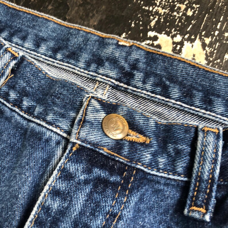 Op de afbeelding: Close-up van een blauwe spijkerbroek. De afbeelding toont de knoop en het knoopsgat, met zichtbare stiksels en een messingkleurige knoop. De jeans heeft een klassieke, versleten uitstraling.