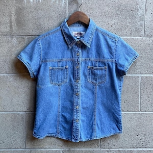 Könnte beinhalten: Ein kurzärmeliges blaues Jeanshemd mit klassischem Kragen, einer durchgehenden Knopfleiste mit Metallknöpfen und zwei Brusttaschen. Das Kleidungsstück verfügt über vertikale Ziernähte und ein Etikett am inneren Halsausschnitt mit der Aufschrift 'L'.