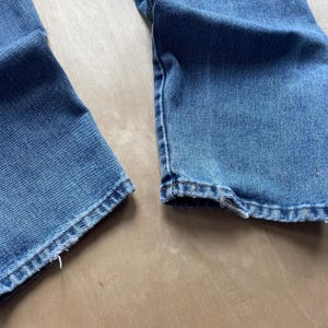 May include: A close-up view of the hem of blue denim jeans. The jeans are a light blue wash, showing the stitching and some fraying. The fabric has a worn appearance, suggesting a vintage style. The jeans are likely made of cotton.