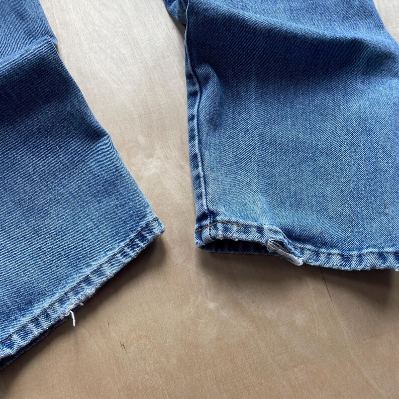May include: A close-up view of the hem of blue denim jeans. The jeans are a light blue wash, showing the stitching and some fraying. The fabric has a worn appearance, suggesting a vintage style. The jeans are likely made of cotton.