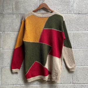 Könnte beinhalten: Ein langärmeliger Strickpullover mit einem farblich abgesetzten Patchwork-Design in Senfgelb, Waldgrün, Dunkelrot und Beige. Die Abschnitte sind durch sichtbare weiße Kreuzstichdetails verbunden, mit einem gerippten Rundhalsausschnitt und Bündchen.