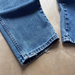 May include: Close-up of the bottom hems of two pairs of blue denim jeans. The jeans are a medium wash, showing signs of wear. The hems are slightly frayed, adding a vintage look.