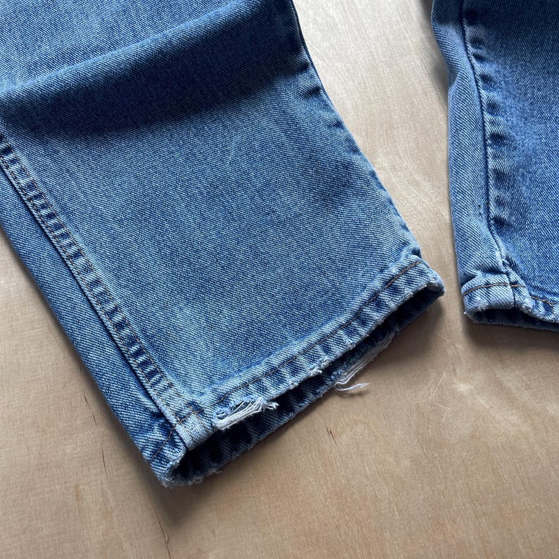May include: Close-up of the bottom hems of two pairs of blue denim jeans. The jeans are a medium wash, showing signs of wear. The hems are slightly frayed, adding a vintage look.