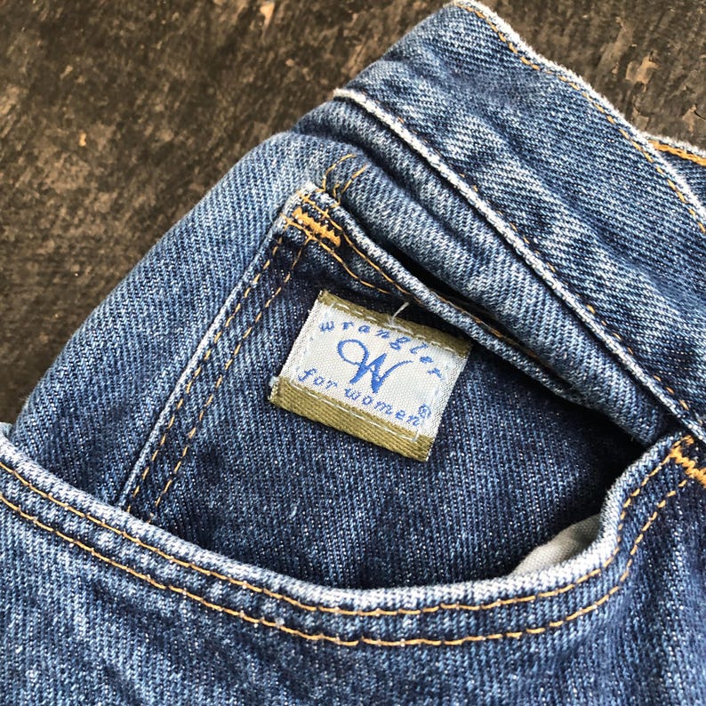 Op de afbeelding: Close-up van een blauwe denim jeans met een zakdetail. De jeans heeft een klein label met de merknaam "Wrangler" en de tekst "for women". De stiksels zijn in een contrasterende gele kleur.