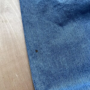 May include: Close-up of blue denim fabric with a small dark spot. The denim has a classic, textured appearance. The fabric is likely from a pair of jeans or a similar garment.
