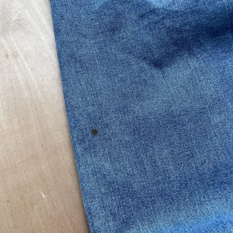 May include: Close-up of blue denim fabric with a small dark spot. The denim has a classic, textured appearance. The fabric is likely from a pair of jeans or a similar garment.