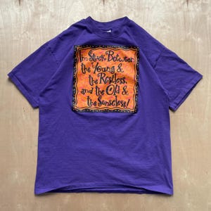 May include: A purple t-shirt with a graphic on the front that reads "I'm Stuck Between the Young & the Restless, and the Old & the Senseless!" The graphic is orange with black and yellow accents.