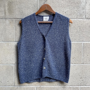 May include: A sleeveless V-neck waistcoat in a speckled navy blue knit fabric. The garment features a front button closure with four light-coloured buttons and a white interior label with the text R&R. The waistcoat is displayed on a wooden hanger.