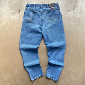 May include: Light blue denim jeans with a classic five-pocket design. The jeans have a straight leg cut and a faded appearance, with a leather patch and a red logo tag on the back. The jeans are made of a durable denim material.