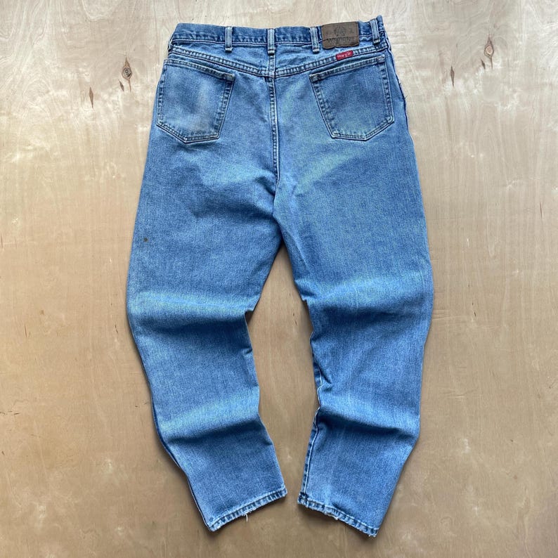 May include: Light blue denim jeans with a classic five-pocket design. The jeans have a straight leg cut and a faded appearance, with a leather patch and a red logo tag on the back. The jeans are made of a durable denim material.