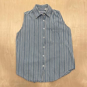 May include: A sleeveless shirt in a blue and white striped design. It features a collared neckline, a single chest pocket, and a curved hem. The fabric looks like a lightweight cotton or blend, ideal for warmer weather. The shirt measures approximately 60 cm in length.