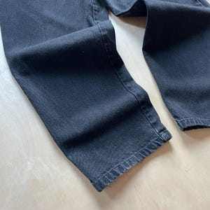 Peut inclure: Gros plan sur un jean en denim noir. Le jean est pli&eacute;, mettant en valeur la texture et les d&eacute;tails des coutures. Le tissu semble &ecirc;tre un denim de poids moyen, avec un aspect l&eacute;g&egrave;rement d&eacute;lav&eacute;. L'image met en &eacute;vidence la jambe et l'ourlet du jean.