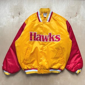 Könnte beinhalten: Eine gelbe Hawks-Jacke aus Satin mit roten Ärmeln. Auf der Jacke steht das Wort "Hawks" in roten Blockbuchstaben auf der Brust. Kragen, Manschetten und Bund sind weiß und rot gestreift.