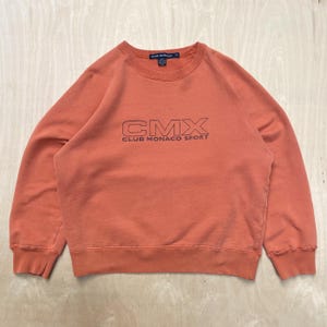 May include: A terracotta-coloured sweatshirt featuring the black text "CMX CLUB MONACO SPORT". The garment has a crew neck and raglan sleeves. The material looks soft and slightly faded.