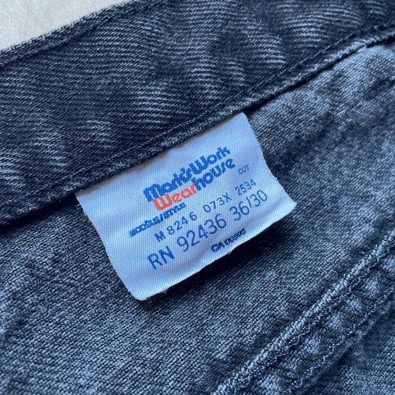 Peut inclure: Gros plan sur une section de jean en denim gris fonc&eacute;. Une &eacute;tiquette blanche avec du texte bleu et orange indique "Mark's Work Wearhouse". L'&eacute;tiquette affiche &eacute;galement la taille 36/30 et d'autres informations de style.