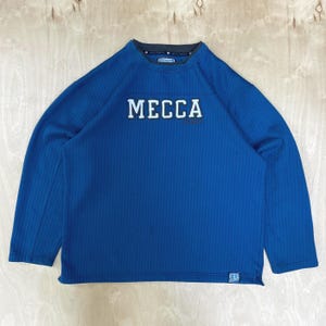 May include: A long-sleeved blue sweatshirt with the word "MECCA" in white block letters across the chest. The sweatshirt has a ribbed texture and a dark gray collar. A small logo is visible on the lower right side.