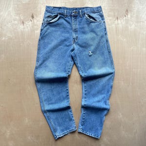 May include: Light blue denim jeans with a classic five-pocket design. The jeans have a faded wash and a distressed area on the right leg. The jeans are laid flat on a light-coloured surface.