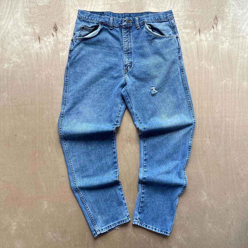 May include: Light blue denim jeans with a classic five-pocket design. The jeans have a faded wash and a distressed area on the right leg. The jeans are laid flat on a light-coloured surface.
