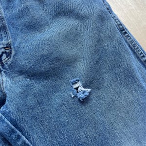 May include: Close-up of blue denim jeans with a distressed, ripped detail. The tear reveals the white threads underneath, adding a worn, vintage aesthetic. The jeans show signs of wear and tear.