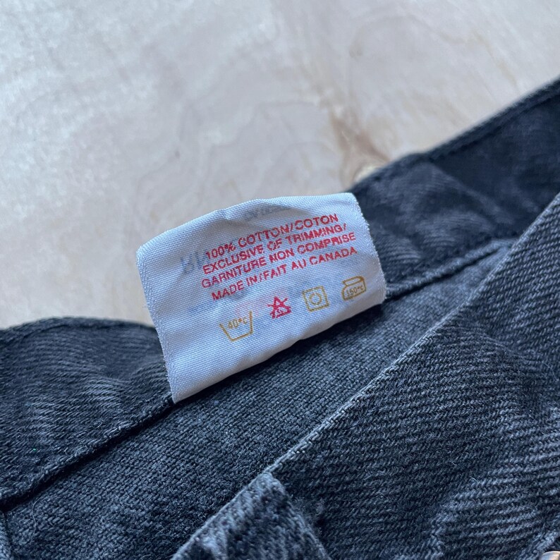 Peut inclure: Gros plan sur une &eacute;tiquette blanche avec du texte noir indiquant "100% COTTON/COTON EXCLUSIVE OF TRIMMING/ GARNITURE MON COMPRISE MADE IN/FAIT AU CANADA". L'&eacute;tiquette est sur un jean en denim gris fonc&eacute;.