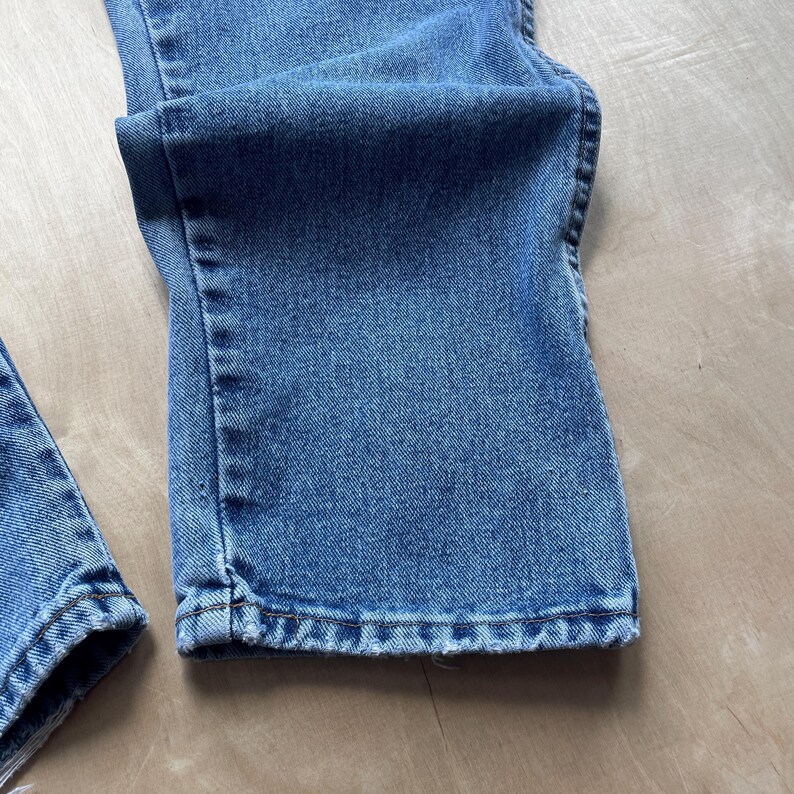 May include: Close-up of light blue denim jeans. The jeans are folded, showing the hem and the fabric's texture. The denim has a classic, slightly faded look, with visible stitching details.