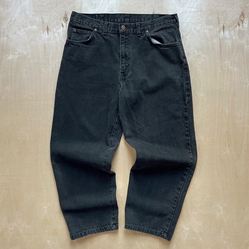 Peut inclure: Un jean en denim noir. Le jean a une conception classique &agrave; cinq poches, une fermeture &agrave; bouton et fermeture &eacute;clair, et des passants de ceinture. Le denim semble &ecirc;tre de poids moyen, avec un aspect l&eacute;g&egrave;rement d&eacute;lav&eacute;. Le jean est pos&eacute; &agrave; plat sur une surface en bois clair.