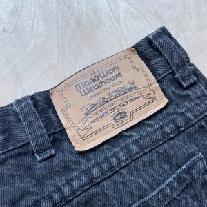 Peut inclure: Gros plan sur un jean en denim noir avec une &eacute;tiquette en cuir. L'&eacute;tiquette indique "Mark's Work Wearhouse" et comprend du texte en anglais et en fran&ccedil;ais. Le jean a une conception classique &agrave; cinq poches.
