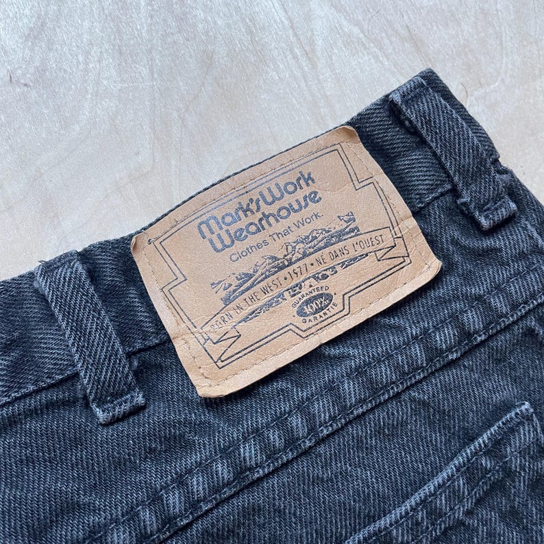 Peut inclure: Gros plan sur un jean en denim noir avec une &eacute;tiquette en cuir. L'&eacute;tiquette indique "Mark's Work Wearhouse" et comprend du texte en anglais et en fran&ccedil;ais. Le jean a une conception classique &agrave; cinq poches.