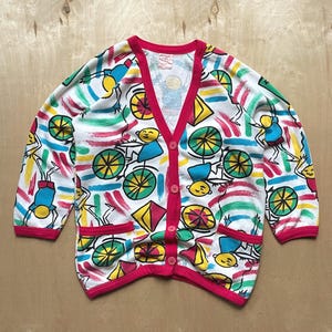 May include: A white cardigan with a colourful pattern of stick figures riding bicycles and geometric shapes. The cardigan has a red trim and a button-up front.