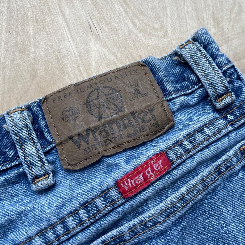 May include: Close-up of blue denim jeans with a brown leather patch and a red tag. The leather patch has the Wrangler Jeans logo and the words "Premium Quality." The red tag has the Wrangler logo.