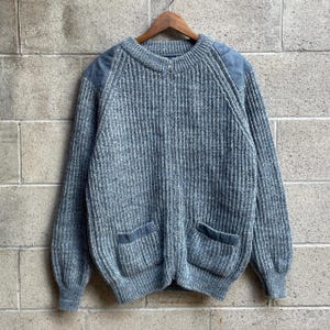 May include: A grey, ribbed knit zip-up sweater featuring a crew neckline, long sleeves, and two front pockets. The garment includes solid grey fabric patches on the shoulders and pocket trim, and it is displayed on a wooden hanger.
