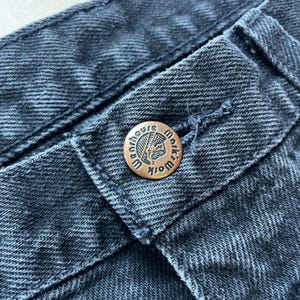 Peut inclure: Gros plan sur un jean en denim fonc&eacute; avec un bouton cuivr&eacute;. Le bouton est grav&eacute; des mots "Warehouse Mark" et d'un profil am&eacute;rindien. Le jean pr&eacute;sente des coutures visibles.