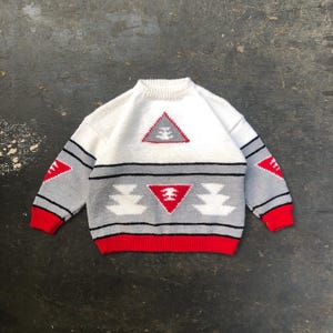 May include: White and grey knitted jumper with a red trim and geometric patterns. The jumper features a large triangle with a smaller triangle inside, and a row of smaller triangles with a larger triangle in the centre.