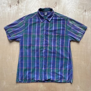 May include: A short-sleeved shirt with a plaid design in blue, green, and pink. The shirt features a chest pocket and the brand name "GAP" is visible on the collar. The shirt has a button-down front.
