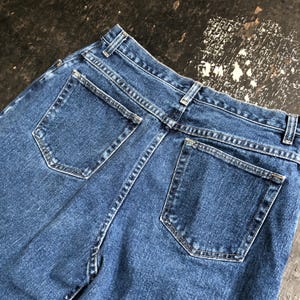 Op de afbeelding: Close-up van de achterkant van een blauwe spijkerbroek. De jeans heeft twee achterzakken, riemlussen en een klassiek ontwerp. De denim heeft een medium wassing en de stiksels zijn zichtbaar. De jeans is waarschijnlijk vintage of pre-owned.