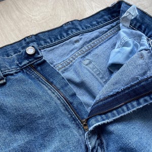 May include: Close-up of light blue denim jeans with an open zip. The jeans have a button closure and visible stitching details. The fabric shows signs of wear, with lighter shades in areas.