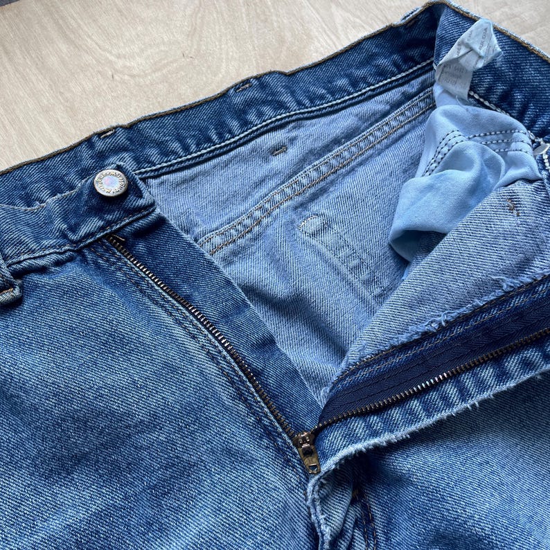 May include: Close-up of light blue denim jeans with an open zip. The jeans have a button closure and visible stitching details. The fabric shows signs of wear, with lighter shades in areas.