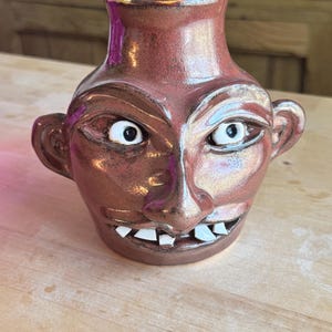 May include: A ceramic vase with a face design. The vase is a reddish-brown color with a metallic sheen. It features a nose, eyes, ears, and a mouth with white teeth. The vase is sitting on a wooden surface.