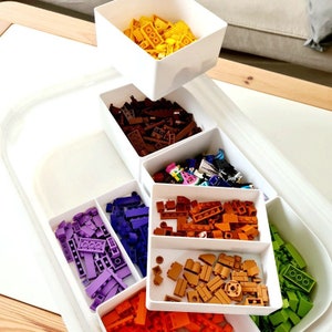 Peut inclure: Un bac de rangement en plastique blanc avec plusieurs compartiments remplis de briques Lego colorées. Les compartiments sont remplis de briques Lego violettes, orange, brunes, vertes, jaunes et beiges.