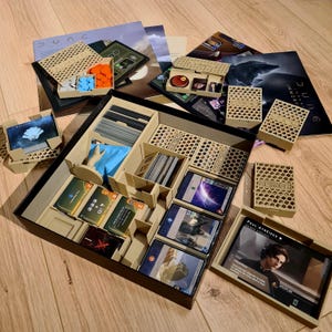 Puede incluir: Un organizador de juego de mesa para el juego Dune: Imperium. El organizador es beige y negro, con varios compartimentos para cartas, fichas y otros componentes del juego. La caja del juego está abierta, revelando el contenido.