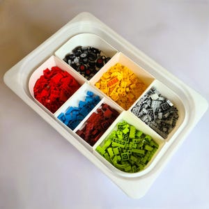 Può includere: Un vassoio di plastica bianco con sei scomparti pieni di mattoncini Lego di diversi colori. Gli scomparti contengono mattoncini Lego rossi, blu, verdi, gialli, grigi e neri.