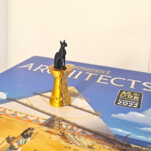 Op de afbeelding: Een zwart kattenbeeldje op een goudkleurige sokkel, geplaatst op een bordspeldoos met de titel "Wonders Architects". De speldoos heeft een blauwe achtergrond en een piramide-illustratie. Het beeldje is een decoratief element.