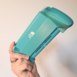 Könnte beinhalten: Eine türkisfarbene Handheld-Spielkonsole mit passendem Schutzgriff. Die Konsole hat ein weißes Logo und einen weißen Akzent auf der Oberseite. Der Griff scheint 3D-gedruckt zu sein und soll den Halt verbessern.