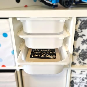 Op de afbeelding: Drie witte plastic opbergbakken gestapeld in een witte kast. De bovenste bak is leeg. De middelste bak is leeg. De onderste bak bevat een bruine kaart met de tekst "Si seulement j'avais plus d'espace" in gouden letters.