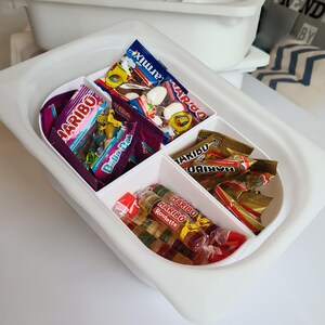 Peut inclure: Un récipient en plastique blanc avec quatre compartiments remplis de différents types de bonbons. Les bonbons comprennent des bonbons gélifiés Haribo, un sac de bonbons gélifiés et un sac de guimauves blanches.