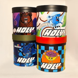 Może przedstawiać: Cztery różne puszki Holy Kombucha, każda z unikalnym wzorem. Puszki są czarne z kolorowymi grafikami. Smaki to Energia, Nawodnienie, Herbata lodowa i Owoc smoka.