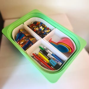 Pode incluir: Um organizador de plástico verde com quatro compartimentos brancos. Os compartimentos contêm canetas, colheres de medição, doces e pequenas figuras de brinquedo. O organizador está sobre uma superfície branca.