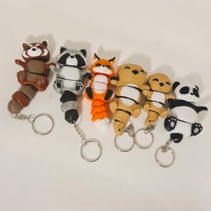 Puede incluir: Una colección de llaveros de animales hechos a mano. Los llaveros presentan un mapache, un zorro, una nutria y un panda. Cada llavero está hecho de arcilla y tiene un llavero plateado adjunto. El mapache es marrón y gris, el zorro es naranja y blanco.